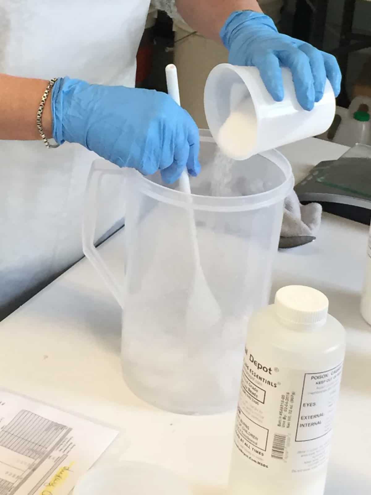 Adding measured sodium hydroxide beads to a pitcher of cold water and stirring to dissolve.
